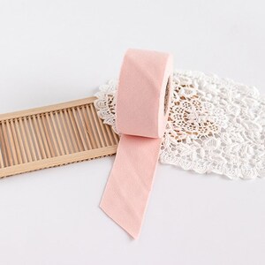 May include: A roll of light pink ribbon with a cut end, unraveled on a white surface. The ribbon is next to a bamboo tray and a white lace doily. The ribbon is a soft, solid color.