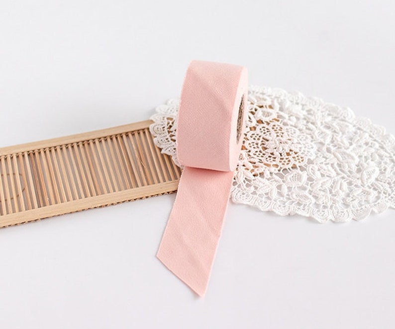 May include: A roll of light pink ribbon with a cut end, unraveled on a white surface. The ribbon is next to a bamboo tray and a white lace doily. The ribbon is a soft, solid color.