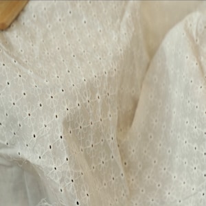 May include: White cotton fabric with a delicate embroidered floral pattern. The fabric has a subtle sheen and a soft, airy feel.