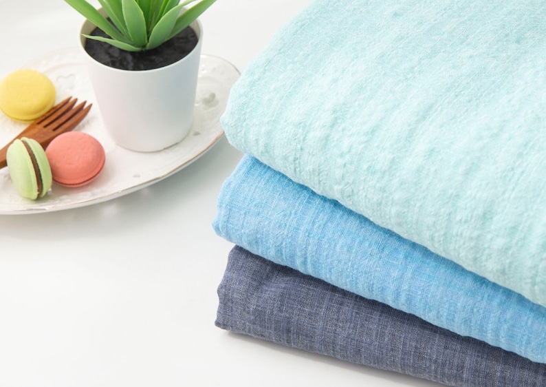 May include: Stack of three fabric swatches in shades of blue. The top swatch is a light blue, the middle swatch is a medium blue, and the bottom swatch is a dark blue. The swatches are folded and arranged on a white surface.
