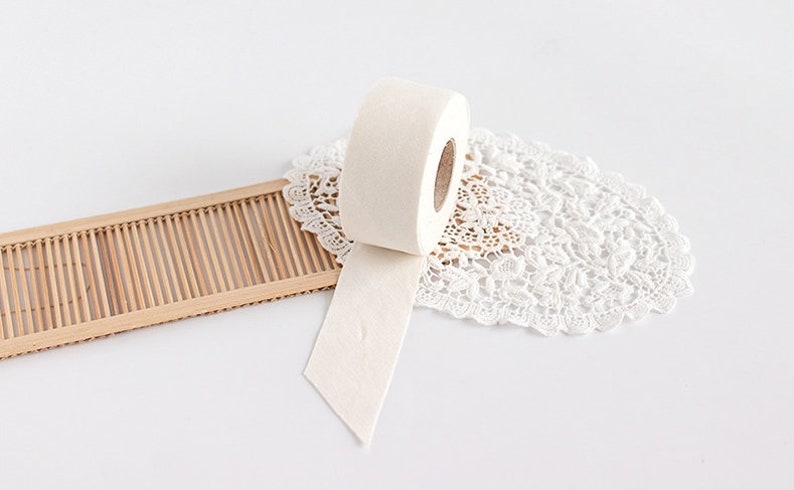 May include: A roll of off-white tape with a textured surface, partially unrolled, resting on a white lace doily and a small bamboo tray. The tape appears to be made of a soft, fibrous material.