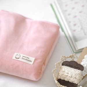 May include: A soft, pink fabric pouch with a small label that reads "Natural style handmade." A decorative basket holds lace trim and a tag that says "fabric story." A book with floral patterns is in the background.