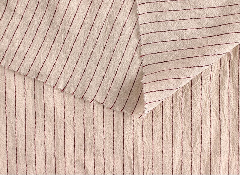 May include: Close-up of a cream-colored fabric with thin, red stripes. The fabric has a textured, slightly rough surface.