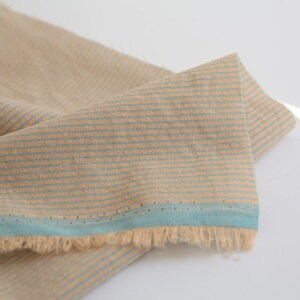 May include: Beige and blue striped fabric with a fringed edge. The fabric has a subtle texture and a light blue trim.