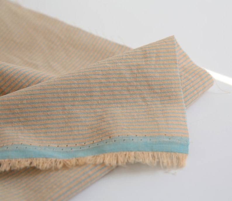 May include: Beige and blue striped fabric with a fringed edge. The fabric has a subtle texture and a light blue trim.