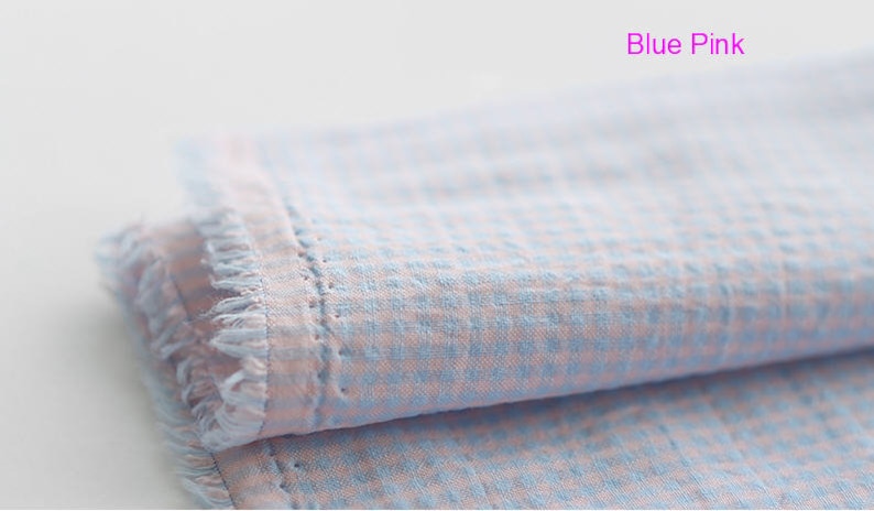 Pu&ograve; includere: Un primo piano di un tessuto gingham blu e rosa con un bordo sfrangiato. Il tessuto &egrave; piegato a met&agrave; e sono visibili i quadrati blu e rosa.