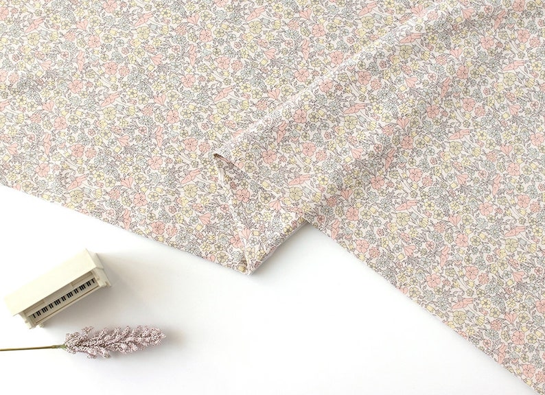 May include: A close-up of a white fabric with a delicate floral pattern in shades of pink, yellow, and grey. The fabric is folded in half, revealing the pattern on both sides.