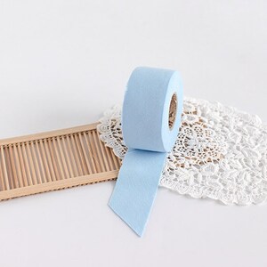 May include: A roll of light blue felt tape with a paper core. The tape is unspooled, revealing a cut edge. The tape sits on a white lace doily and a bamboo tray, against a white background.