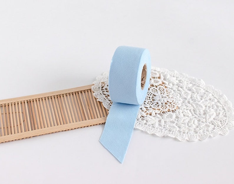 May include: A roll of light blue felt tape with a paper core. The tape is unspooled, revealing a cut edge. The tape sits on a white lace doily and a bamboo tray, against a white background.