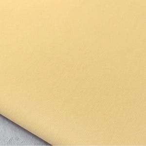 Puede incluir: Primer plano de una tela amarillo pálido. El material tiene un tejido texturizado y está doblado, revelando un vistazo de una superficie gris debajo. El color de la tela es un amarillo suave y apagado.