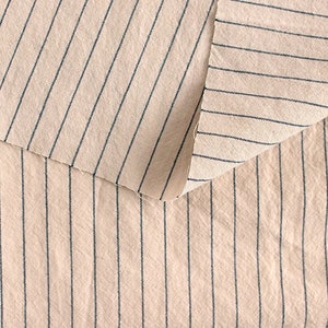 May include: Close-up of a beige fabric with thin black stripes. The fabric is folded over itself, showing the same pattern on both sides.