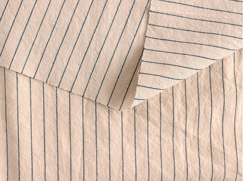 May include: Close-up of a beige fabric with thin black stripes. The fabric is folded over itself, showing the same pattern on both sides.