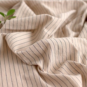 May include: Close-up of a beige fabric with thin black stripes. The fabric is wrinkled and has a soft, natural texture.