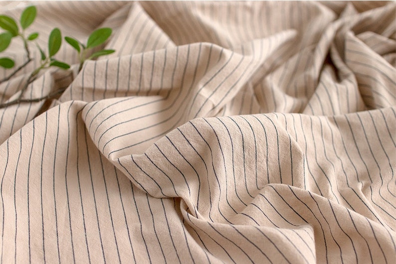 May include: Close-up of a beige fabric with thin black stripes. The fabric is wrinkled and has a soft, natural texture.