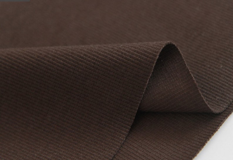May include: Close-up of a brown corduroy fabric with a subtle ribbed texture. The fabric is folded in half, showing the soft, plush surface.
