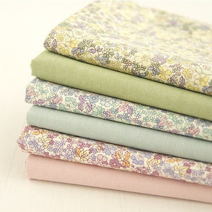 May include: A stack of six fabric swatches featuring floral patterns in shades of green, blue, and pink. The fabrics are arranged in a graduated colour scheme, with the greenest fabric at the bottom and the pinkest fabric at the top.