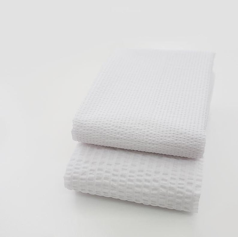 May include: Two white waffle weave kitchen towels stacked on top of each other. The towels are folded in half and have a soft, textured surface.