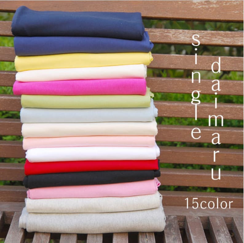 May include: A stack of 15 different colored fabric swatches, including navy blue, yellow, white, pink, green, light blue, beige, peach, red, black, light pink, and gray. The fabric swatches are arranged on a wooden bench with the text "single daimar u 15color" visible on the right side of the image.