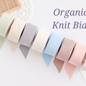 May include: A selection of colorful organic knit bias tape rolls in pastel shades, including green, beige, lavender, white, blue, brown, and pink. The text "Organic Knit Bias" is displayed in purple.