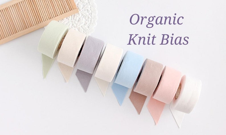 May include: A selection of colorful organic knit bias tape rolls in pastel shades, including green, beige, lavender, white, blue, brown, and pink. The text "Organic Knit Bias" is displayed in purple.