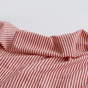 May include: A close-up shot of a red and white striped fabric. The material is folded and draped, revealing the vertical stripe pattern. The fabric looks lightweight, possibly cotton or linen, with a soft texture.