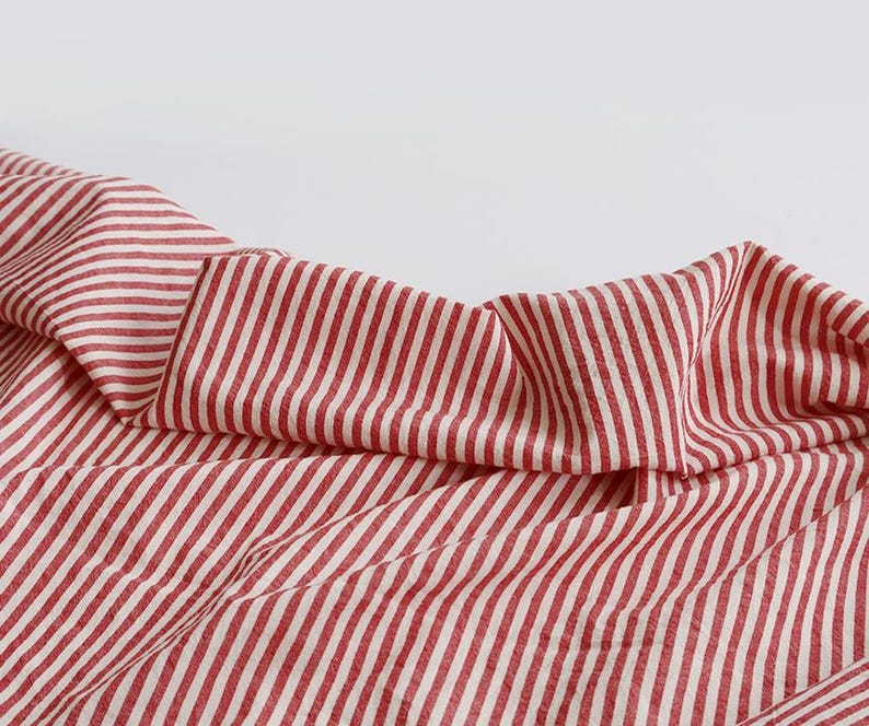 May include: A close-up shot of a red and white striped fabric. The material is folded and draped, revealing the vertical stripe pattern. The fabric looks lightweight, possibly cotton or linen, with a soft texture.