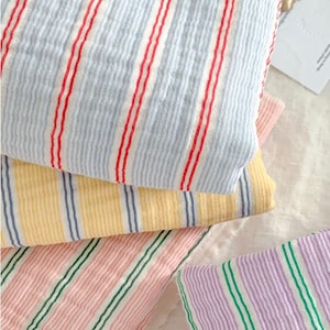 May include: Close-up of folded fabric in various colors with vertical stripes. The fabrics include light blue with red stripes, yellow with blue stripes, pink with green stripes, and purple with green stripes. The fabric appears to be a lightweight cotton or linen.