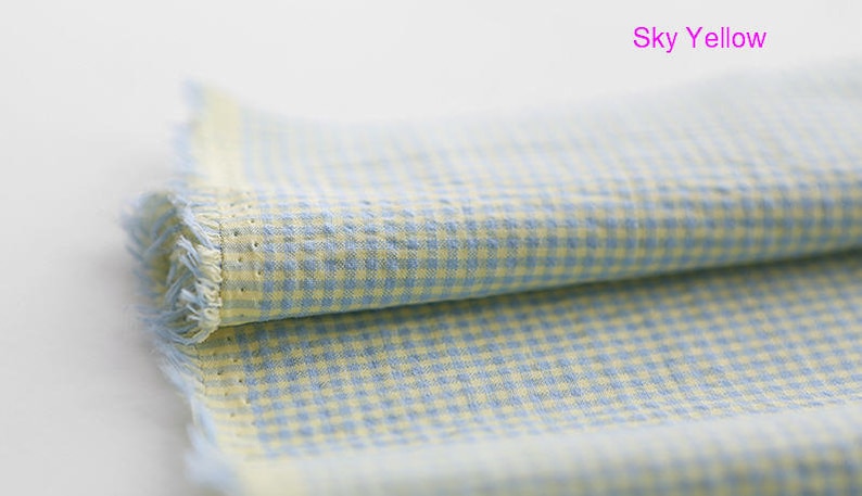 Pu&ograve; includere: Un pezzo di stoffa piegato con un motivo a quadri blu e gialli. La stoffa ha un bordo sfrangiato e il testo "Sky Yellow" &egrave; scritto in rosa sopra la stoffa.