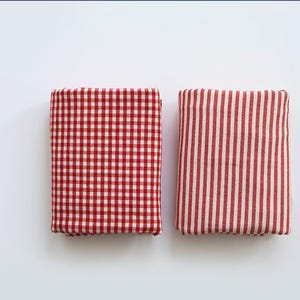 May include: Two folded fabric squares. One has a red and white gingham pattern, the other has red and white vertical stripes. Both are rectangular and appear to be made of cotton or a similar material. The fabrics are neatly folded.