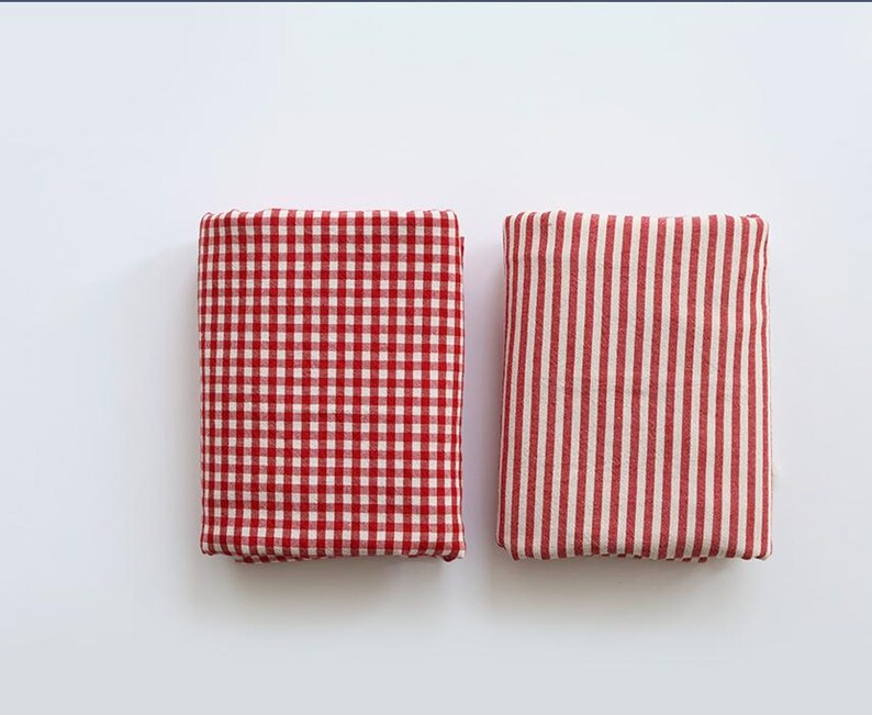 May include: Two folded fabric squares. One has a red and white gingham pattern, the other has red and white vertical stripes. Both are rectangular and appear to be made of cotton or a similar material. The fabrics are neatly folded.