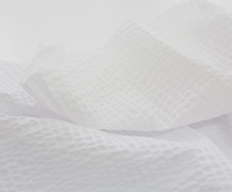 May include: White waffle weave fabric with a textured surface. The fabric is folded and draped, creating soft folds and curves.