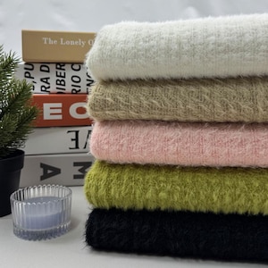 Puede incluir: Una pila de tela suave y texturizada en varios colores: crema, beige, rosa claro, verde oliva y negro. La tela parece suave y afelpada, adecuada para proyectos de ropa o decoración del hogar. The Lonely Of es visible en un libro.