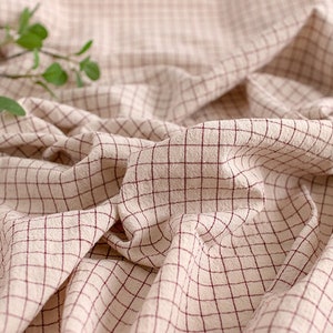 May include: A close-up of a beige fabric with a red checkered pattern. The fabric is folded and wrinkled, creating a textured surface.