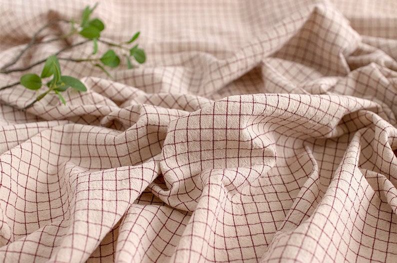 May include: A close-up of a beige fabric with a red checkered pattern. The fabric is folded and wrinkled, creating a textured surface.
