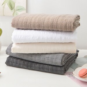 May include: A stack of four fabric swatches in different colors and textures. The swatches are folded and stacked on a white surface. The top swatch is a light brown color, the second swatch is white, the third swatch is a light beige color, and the bottom swatch is a dark gray color.