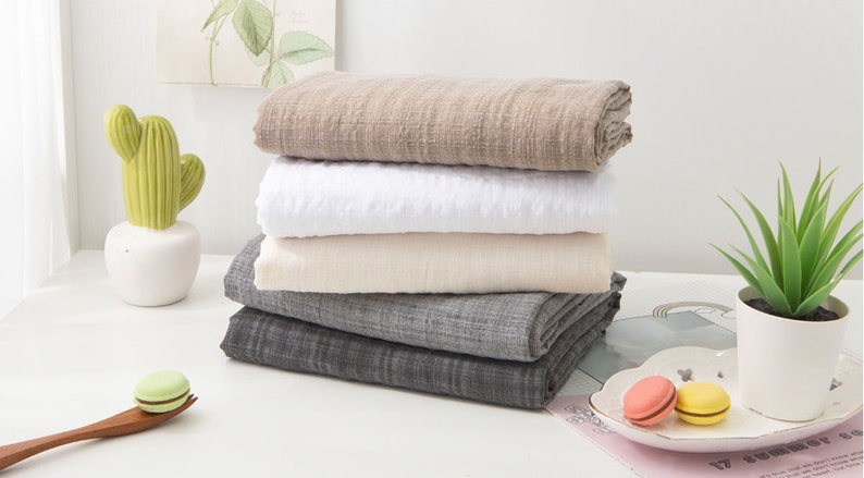 May include: A stack of four fabric swatches in different colors and textures. The swatches are folded and stacked on a white surface. The top swatch is a light brown color, the second swatch is white, the third swatch is a light beige color, and the bottom swatch is a dark gray color.