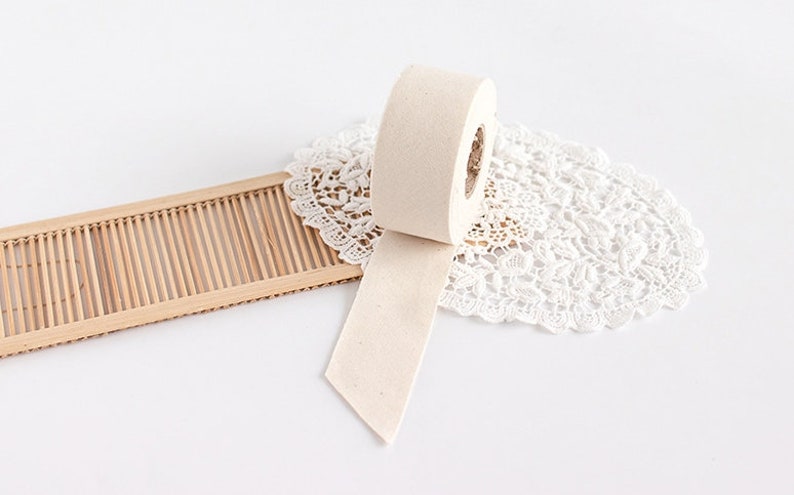 May include: A roll of beige masking tape with a partially unwound strip, resting on a white lace doily. A small, light brown bamboo tray is in the background. The tape roll is positioned at an angle, with the end of the tape pointing downwards.