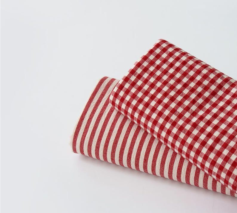 May include: Two folded fabric pieces. One is red and white gingham, the other is red and white striped. The fabrics are layered on a white background. The fabrics are likely for home decor or crafting.