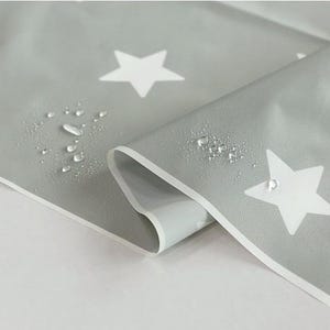 Puede incluir: Tela gris impermeable con un patrón de estrellas blancas. Se ven gotas de agua en la superficie. La tela está doblada, mostrando un reverso blanco. Adecuada para diversos proyectos de manualidades.