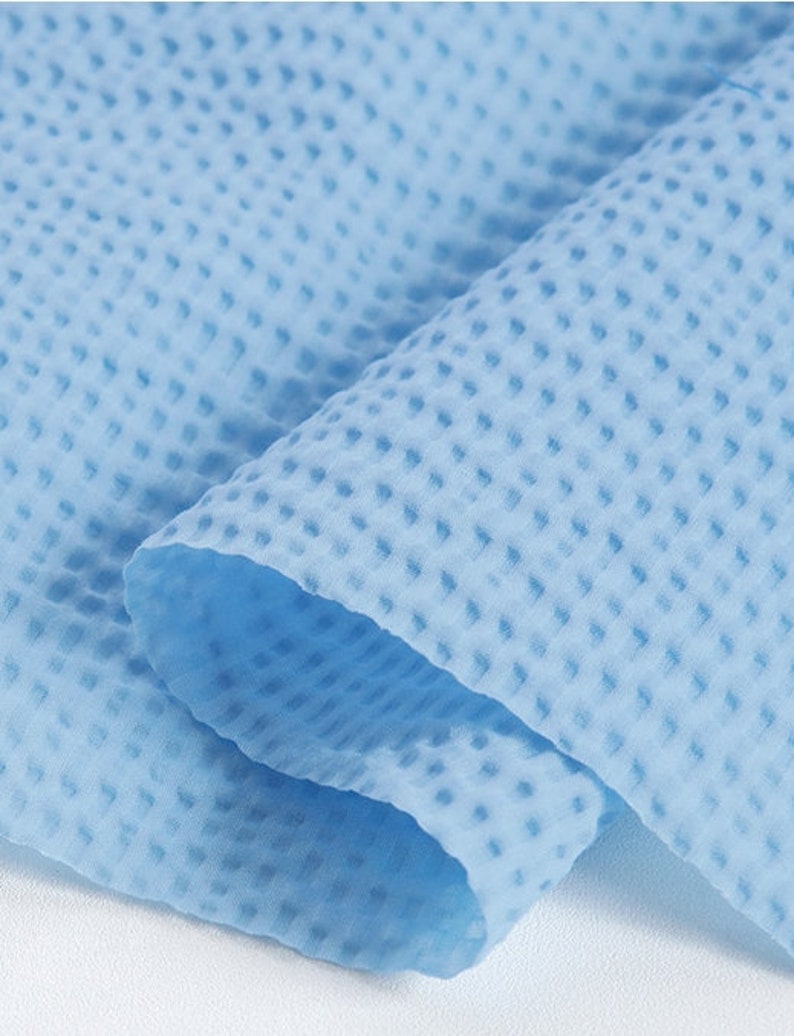 May include: Close-up of a light blue, textured fabric with a waffle-like pattern. The fabric is folded, revealing layers and shadows. The material appears soft and lightweight, suitable for various applications.