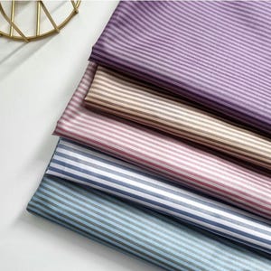 May include: A stack of fabric samples in various colors, including purple, tan, pink, and blue, each with a striped pattern. The fabrics are neatly folded, showcasing the different color options and textures.