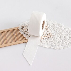 May include: A roll of white, textured paper with a cut edge, resting on a white lace doily. A small, light brown bamboo tray is to the left. The background is white.