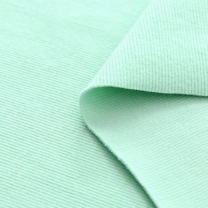 Puede incluir: Primer plano de una sección de tela verde menta. La tela tiene una textura acanalada, con líneas horizontales visibles que recorren la superficie. Una porción de la tela está doblada, revelando la parte inferior y agregando profundidad a la imagen.