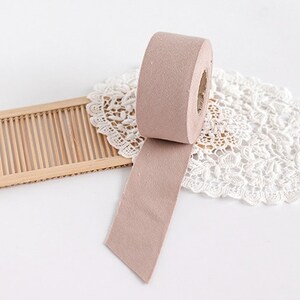 May include: A roll of light brown felt tape with a section unrolled, resting on a white lace doily. A small, rectangular wooden mat is to the left of the tape. The background is white.