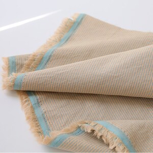 May include: A beige fabric with blue stripes and a frayed edge. The fabric is folded in half and the blue stripes are visible on both sides.