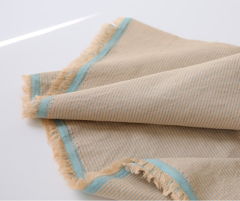May include: A beige fabric with blue stripes and a frayed edge. The fabric is folded in half and the blue stripes are visible on both sides.