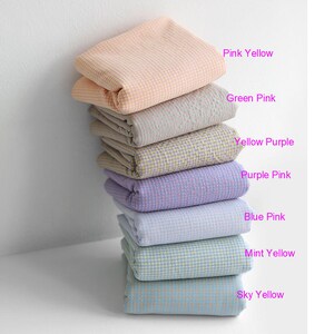 Pu&ograve; includere: Una pila di otto quadrati di tessuto piegati in vari colori pastello, tra cui rosa, giallo, verde, viola, blu e menta. Ogni quadrato ha un piccolo motivo a scacchi.