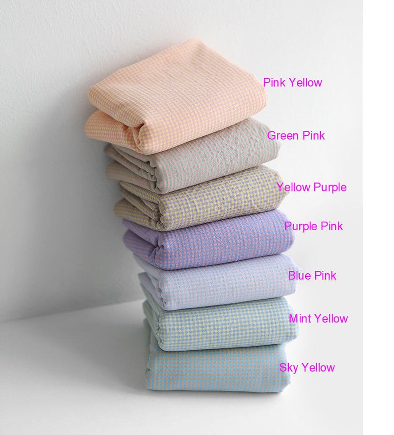 Pu&ograve; includere: Una pila di otto quadrati di tessuto piegati in vari colori pastello, tra cui rosa, giallo, verde, viola, blu e menta. Ogni quadrato ha un piccolo motivo a scacchi.