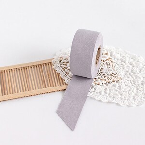 May include: A roll of light purple washi tape with a cut end, unspooled on a white surface. The tape is next to a small, rectangular bamboo tray and a white lace doily.