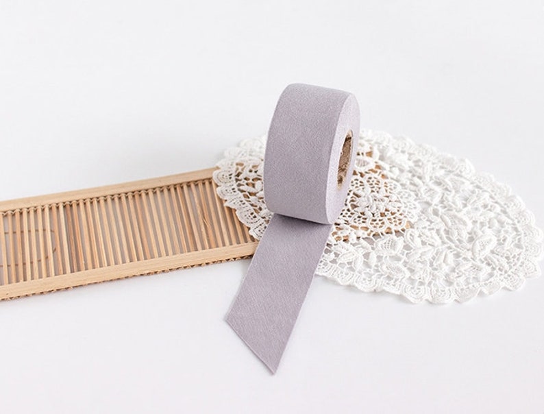 May include: A roll of light purple washi tape with a cut end, unspooled on a white surface. The tape is next to a small, rectangular bamboo tray and a white lace doily.